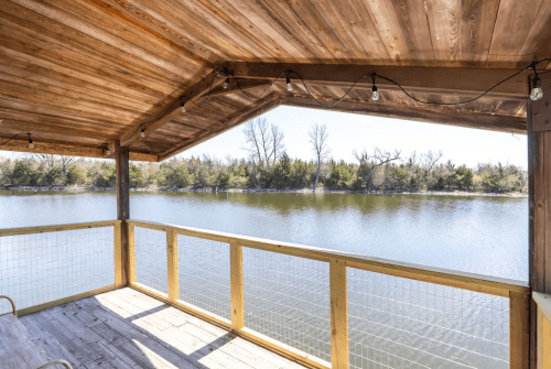 A wooden deck overlooking a calm lake, surrounded by trees and a clear blue sky. String lights hang above.
