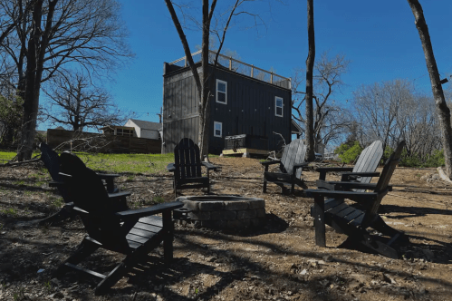 A cozy outdoor space with four black chairs around a fire pit, set against a backdrop of a wooden house and trees.