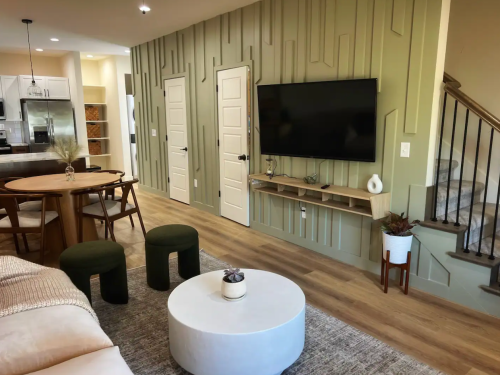Modern living room featuring a green accent wall, a round coffee table, dining table, and a large TV.