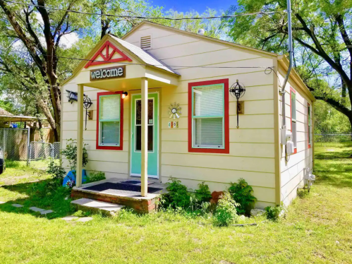 A charming yellow house with red accents, a welcoming sign, and a green door, surrounded by greenery.