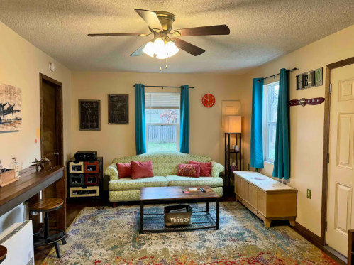 Cozy living room with a green patterned couch, wooden coffee table, and colorful decor, featuring a ceiling fan and bright curtains.