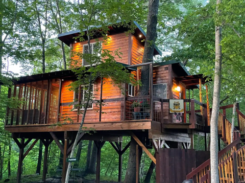 A wooden treehouse nestled among trees, featuring multiple levels and large windows, illuminated in the evening.