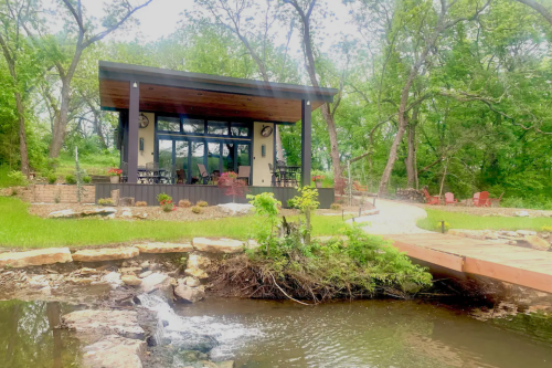 A cozy cabin surrounded by greenery, with a small stream and a wooden bridge leading to a patio area.