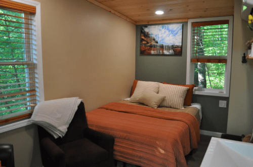 Cozy bedroom with a bed, orange blanket, and a chair, surrounded by windows and natural light.
