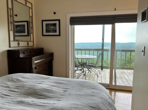 Cozy bedroom with a wooden dresser, mirror, and a view of a balcony overlooking a green landscape.