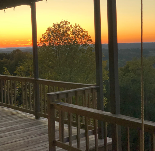 A wooden deck overlooks a scenic sunset with vibrant orange and yellow hues, surrounded by trees and rolling hills.
