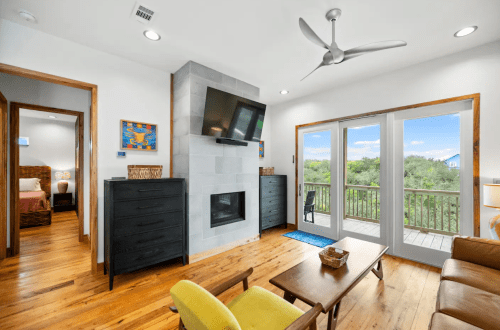 Cozy living area with a fireplace, TV, and sliding doors to a balcony overlooking greenery. Modern decor and wooden floors.