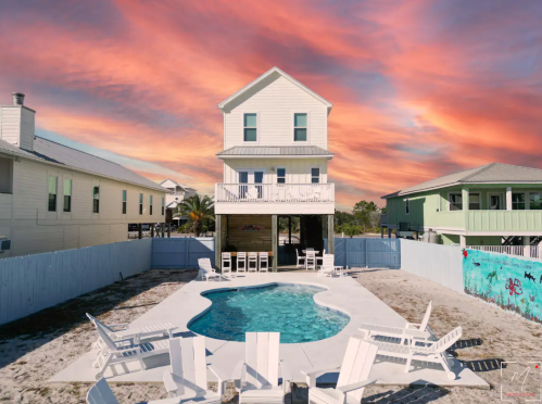 A beach house with a pool surrounded by white chairs, set against a vibrant sunset sky.