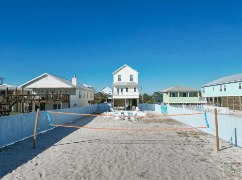 A beach house surrounded by sand, with a volleyball net set up in the foreground and other houses in the background.