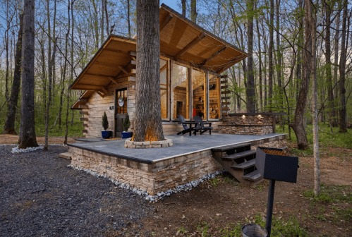 A modern cabin with large windows, surrounded by trees, featuring a stone patio and outdoor seating area.