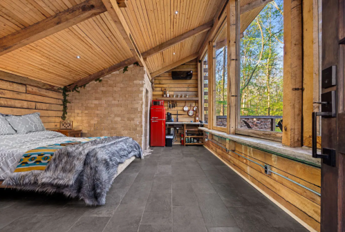 Cozy cabin interior with a wooden ceiling, large windows, a bed, and a red fridge, surrounded by nature.