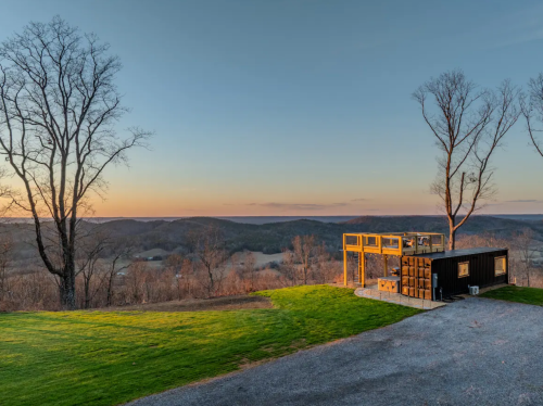 A modern cabin with a glass deck overlooks a scenic valley at sunset, surrounded by bare trees and rolling hills.