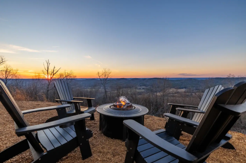 Four black chairs surround a fire pit, overlooking a scenic sunset and rolling hills in the background.