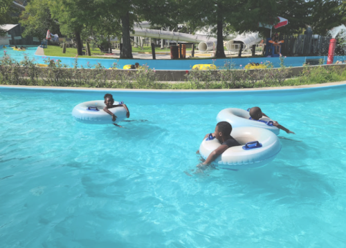 Three children float on inner tubes in a blue pool, surrounded by greenery and water slides in the background.
