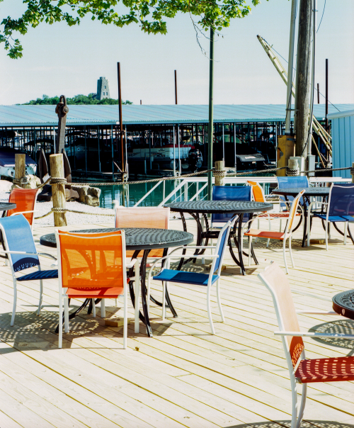 Colorful chairs and tables on a wooden deck by a marina, with a boat dock and a distant lighthouse in the background.