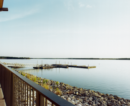 A serene view of a calm lake with a wooden dock and rocky shoreline under a clear blue sky.