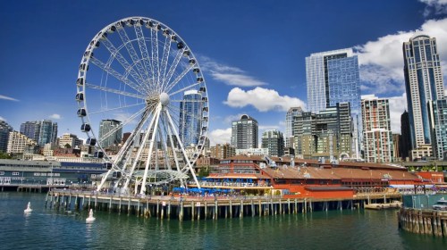A large Ferris wheel stands by the waterfront, surrounded by modern buildings and a clear blue sky.
