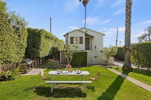 A cozy backyard with a dining table, chairs, and a grill, surrounded by lush greenery and a charming house.