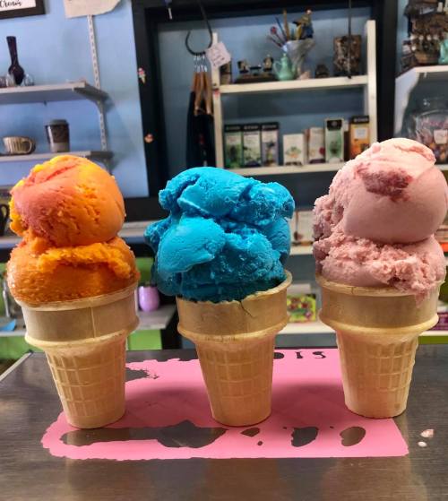 Three ice cream cones with vibrant scoops: orange, blue, and pink, displayed on a counter in a colorful shop.
