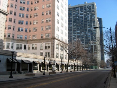 A city street lined with tall buildings, featuring a mix of modern and older architecture, under a clear blue sky.