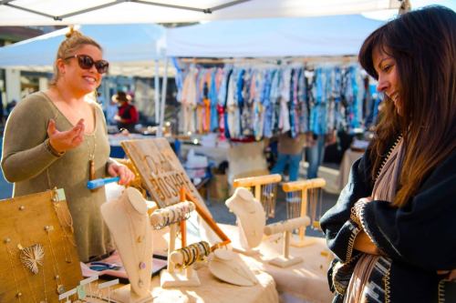 A vendor shows jewelry to a customer at an outdoor market, with colorful displays in the background.