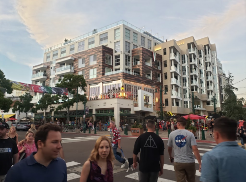 A busy street scene with people walking, featuring modern buildings and outdoor dining areas under a cloudy sky.