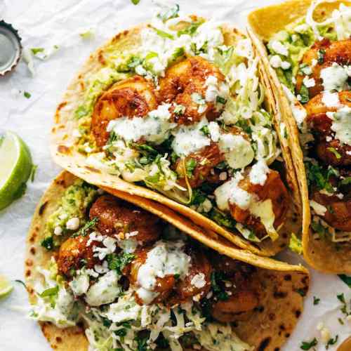 Three tacos filled with shrimp, guacamole, cabbage, and drizzled with creamy sauce, garnished with cilantro and lime.