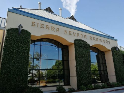 Exterior of Sierra Nevada Brewery, featuring large windows and greenery, under a clear blue sky.