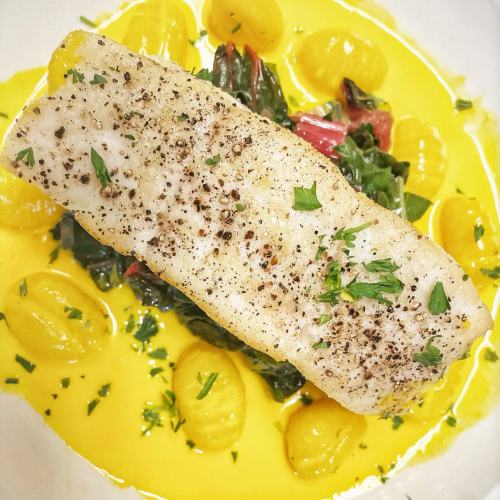 A fillet of fish on a bed of greens and yellow sauce, surrounded by small gnocchi, garnished with herbs.
