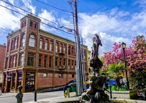 A historic brick building with large windows, a statue in the foreground, and blooming trees under a blue sky.