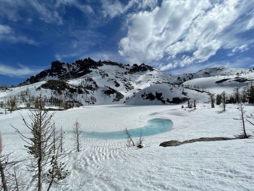 A snowy landscape with a frozen lake, surrounded by mountains and scattered trees under a blue sky with clouds.