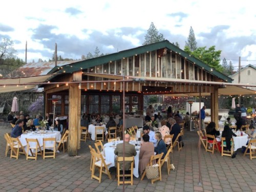 Outdoor dining scene at a rustic venue, with guests seated at tables under string lights and a cloudy sky.