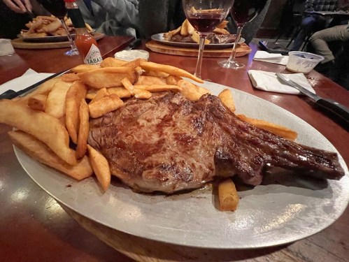 A large ribeye steak served with golden fries on a metal platter, accompanied by glasses of red wine.
