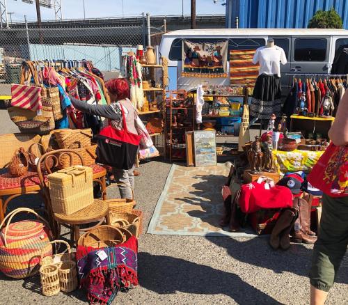 A vibrant outdoor market scene with various handmade crafts, bags, and vintage items displayed for sale.
