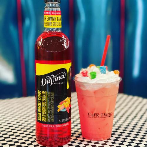 A bottle of DaVinci Gourmet sour gummy candy syrup next to a pink drink topped with whipped cream and colorful candy.