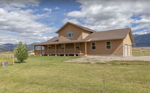A large, tan farmhouse with a porch, set against a backdrop of mountains and a grassy field under a cloudy sky.