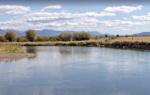 A serene river flows through a grassy landscape, with mountains and fluffy clouds in the background.
