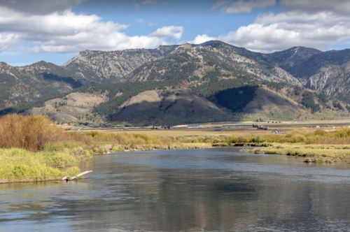 A serene river flows through a valley surrounded by mountains and lush greenery under a partly cloudy sky.