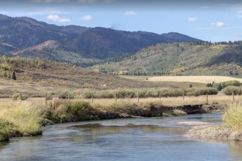 A serene river flows through a valley surrounded by rolling hills and mountains, with colorful autumn foliage.