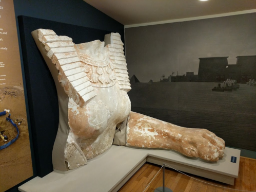 A large ancient statue fragment featuring a torso with decorative elements and a lion's paw, displayed in a museum.