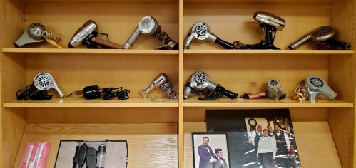 A wooden shelf displaying various vintage hair dryers and a framed photo of people in formal attire.