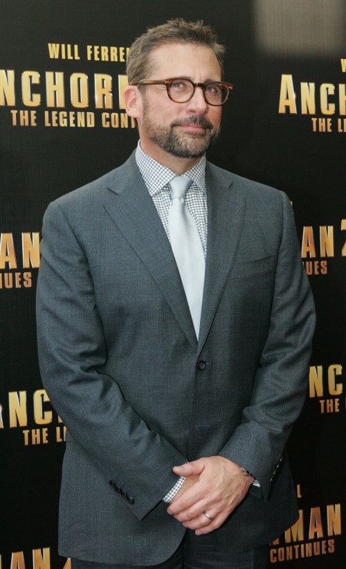 A man in a gray suit and glasses stands confidently at a movie premiere, smiling with arms crossed.