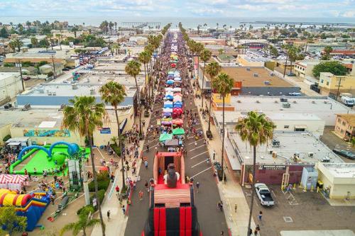 Aerial view of a vibrant street festival with colorful tents, palm trees, and crowds enjoying activities along the coast.