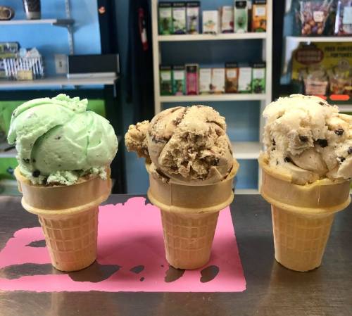 Three ice cream cones with mint green, chocolate chip cookie dough, and vanilla flavors on a pink surface.