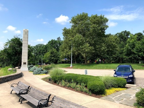 A park scene featuring a tall monument, benches, greenery, and a parked blue car under a clear sky.
