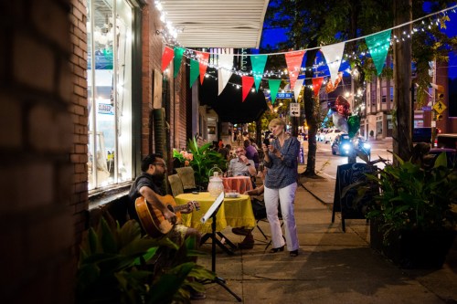 A musician plays guitar outside a café, while a woman records the performance amidst festive decorations and lights.