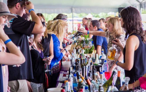 A lively crowd at a beverage tasting event, with people sampling drinks at a long table filled with various bottles.