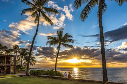 A serene beach scene at sunset, with palm trees silhouetted against colorful clouds and a couple sitting by the water.