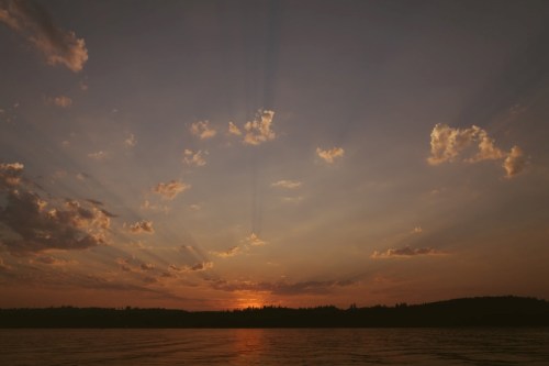 A serene sunset over a calm lake, with rays of light breaking through scattered clouds and a silhouette of trees in the distance.