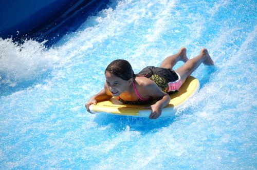 A child rides a boogie board down a water slide, splashing through bright blue water with a joyful expression.